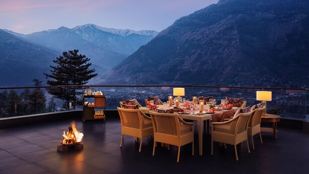 A dining area with a table set for four, surrounded by wicker chairs, a fire feature, and a view of mountains. - Baragarh Resort & Spa, Manali- IHCL SeleQtions (Kullu)