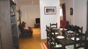 Uma área de refeições com uma mesa de madeira posta para quatro pessoas, rodeada por cadeiras. Há uma televisão ao fundo, e uma cadeira vermelha é visível lateralmente.