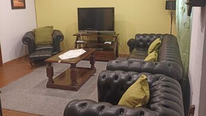 Uma sala de estar com sofá de couro preto, mesa de centro de madeira, televisão em um móvel e um aparelho de ar condicionado na parede.