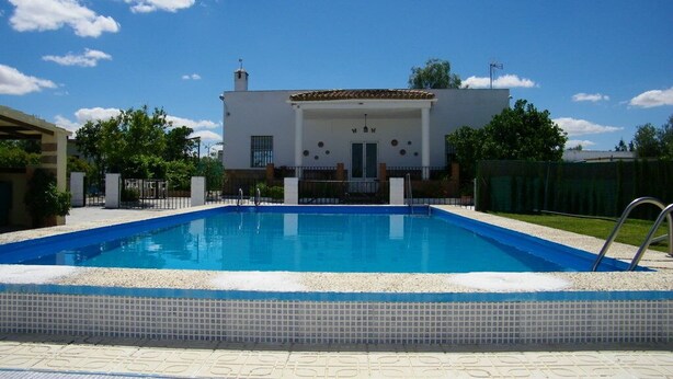 Piscina com deck ladrilhado, edifício branco com telhado em azulejos e céu azul límpido.