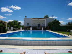 Une piscine avec une terrasse carrelée, un bâtiment blanc doté d’un toit en tuiles et un ciel d’un bleu limpide.