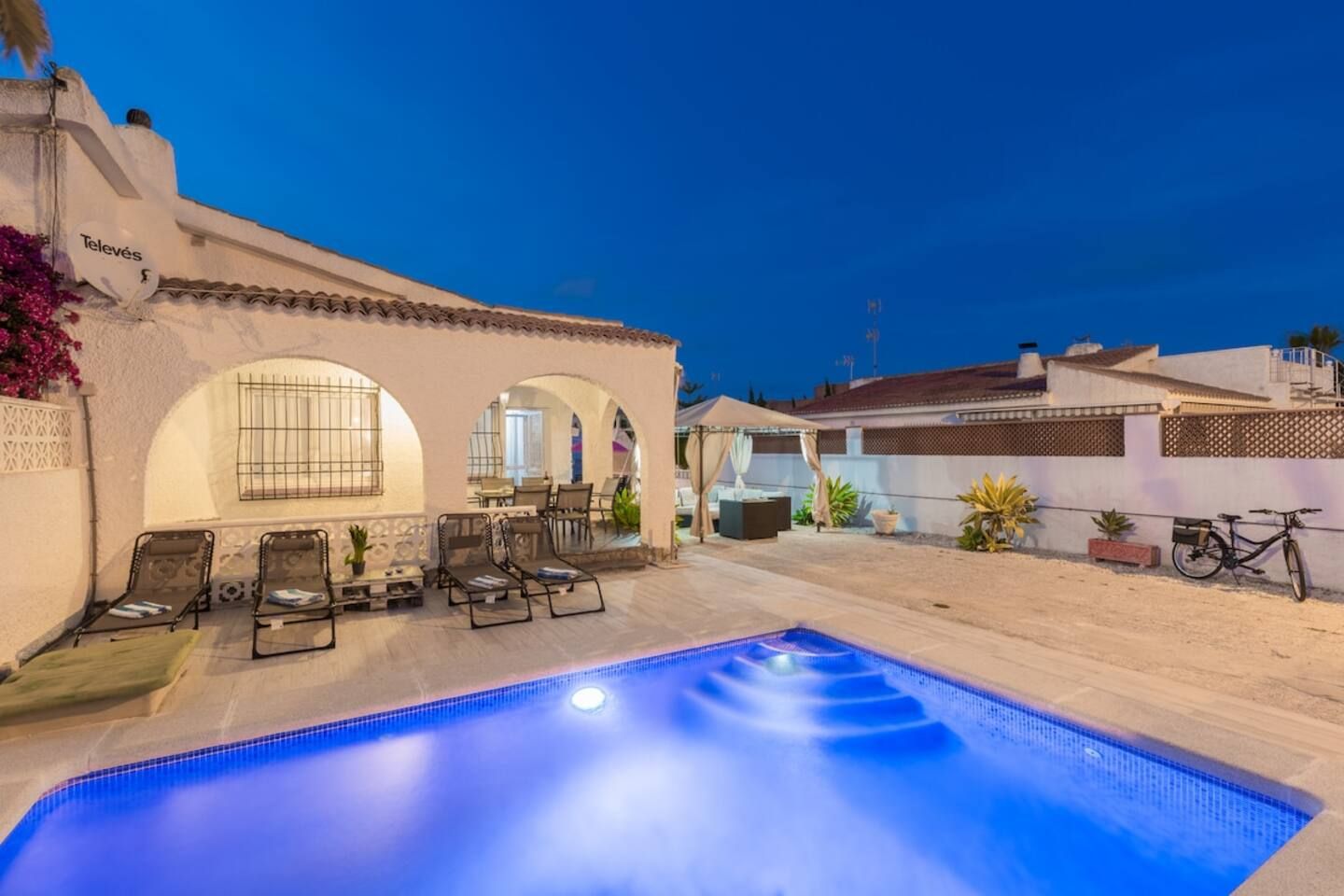 Una piscina con sillas de descanso y una zona de estar cubierta, rodeada por un edificio blanco con ventanas de arco.