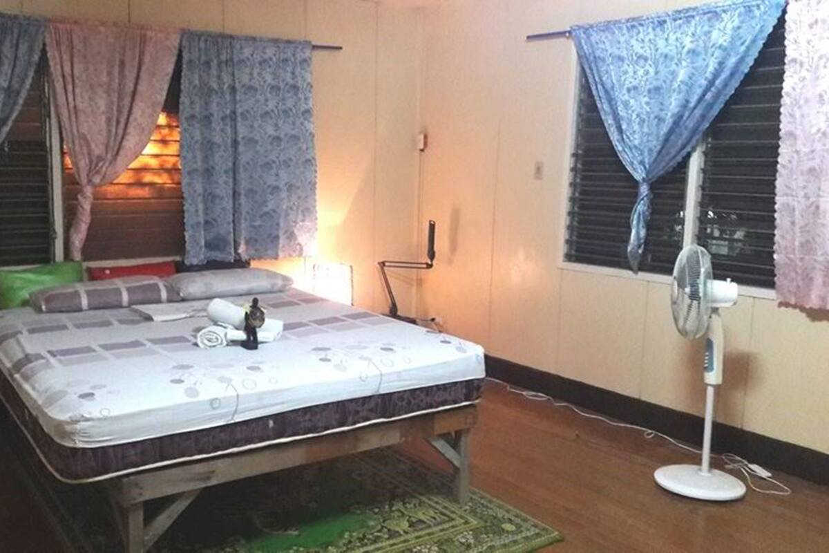 Une chambre avec un lit simple, un ventilateur sur pied et une fenêtre avec des rideaux.