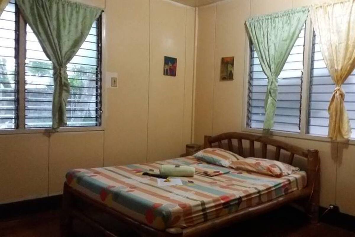 Une chambre avec un lit simple, deux fenêtres avec des rideaux et une petite table de chevet.