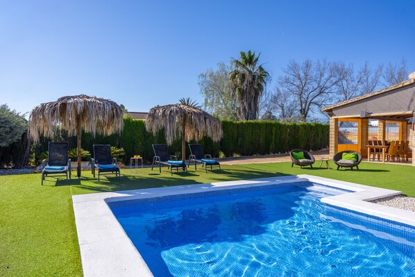 A swimming pool with lounge chairs and umbrellas, surrounded by a well-maintained lawn and a building with a tiled roof.