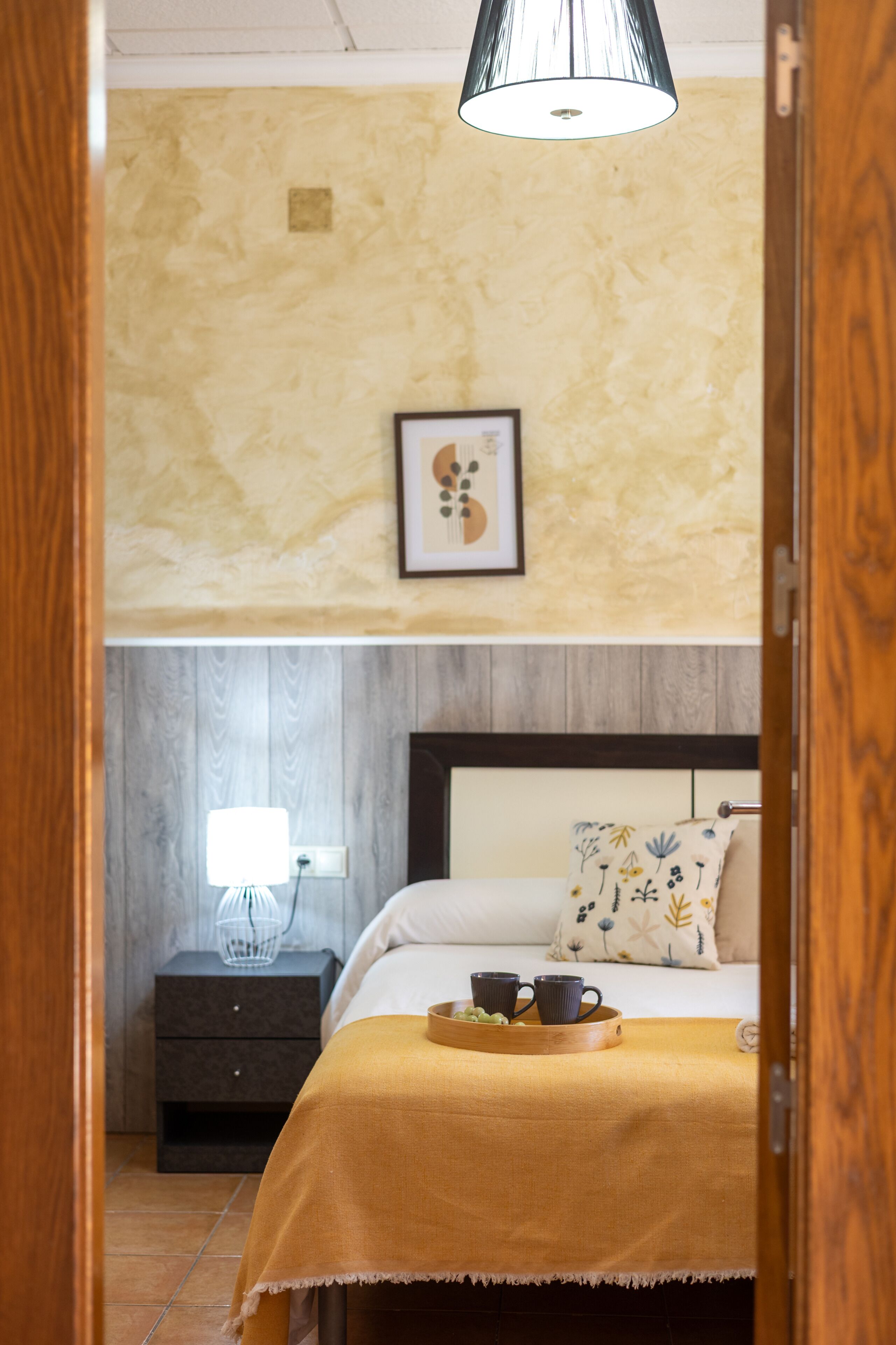 Un dormitorio con cama, mesita de noche, lámpara y un cuadro en la pared.
