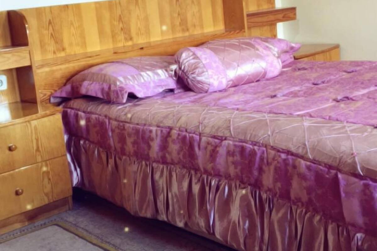 A bedroom with a wooden headboard, a purple bedspread, and a floral-patterned rug.