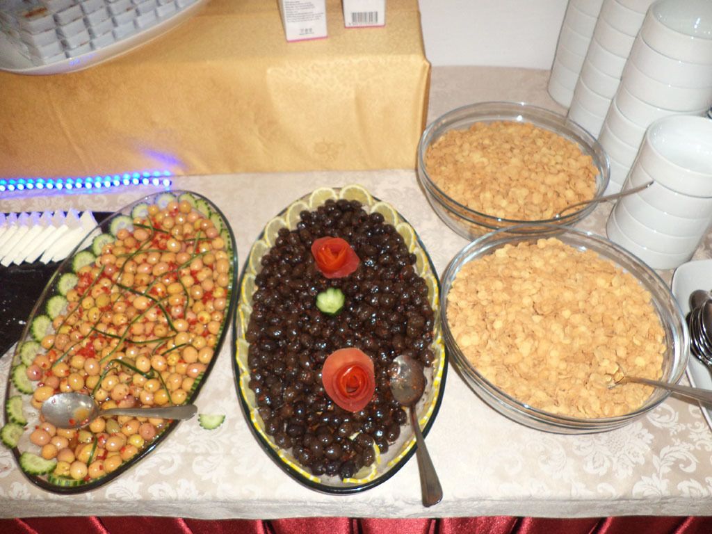 Una mesa de buffet con diversos platos, entre ellos una ensalada con pepinos y tomates cherry, un plato con frijoles negros y una decoración en forma de rosa, y un tazón de cereales.