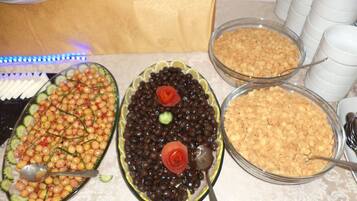 Una mesa de buffet con diversos platos, entre ellos una ensalada con pepinos y tomates cherry, un plato con frijoles negros y una decoración en forma de rosa, y un tazón de cereales.