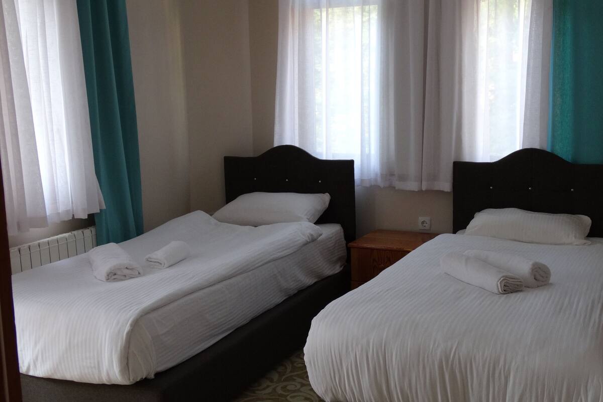 Two single beds with white linens, a wooden nightstand, and a window with sheer curtains.