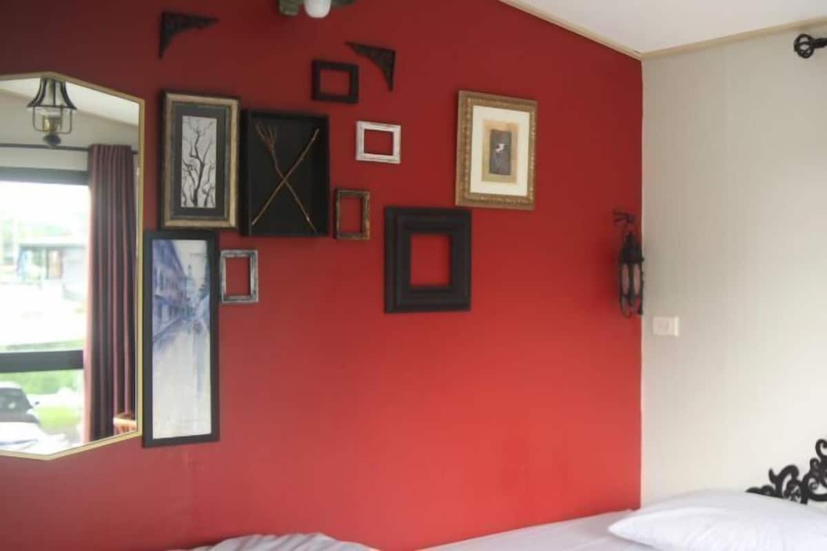 A room with a red wall, a bed with white bedding, and various framed pictures and mirrors on the wall.
