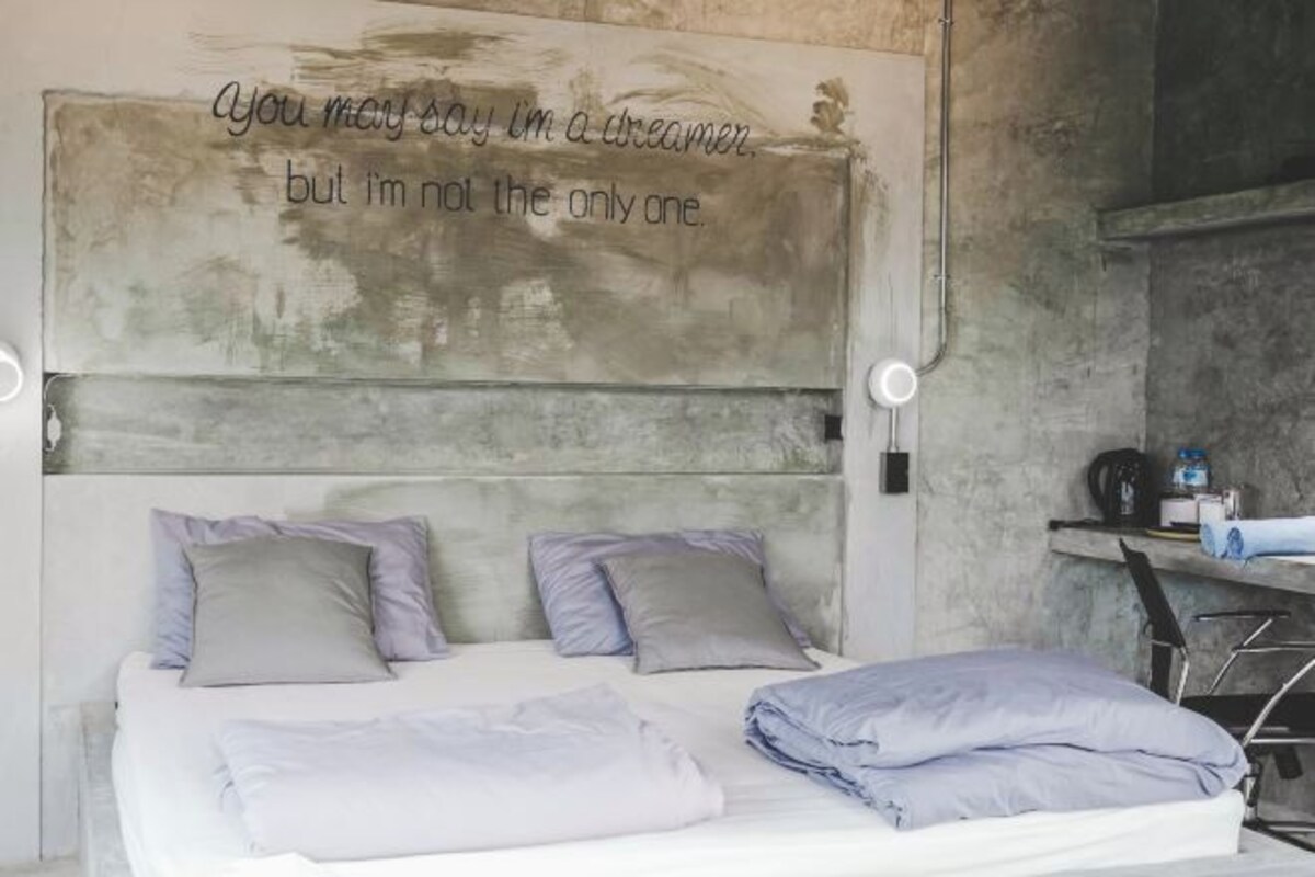 A bedroom with a bed, pillows, a headboard with a motivational quote, and a small desk with a lamp.