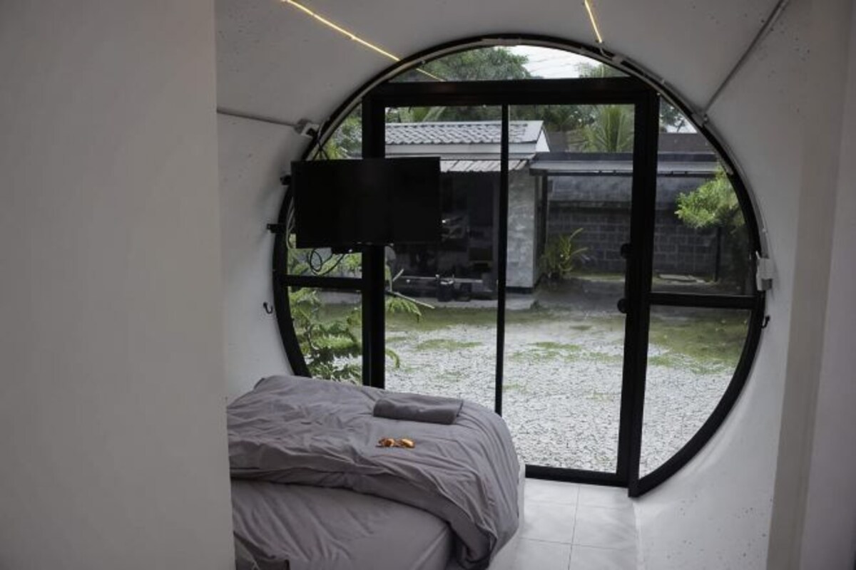 A bedroom with a bed, a TV mounted on the wall, and a large circular window.