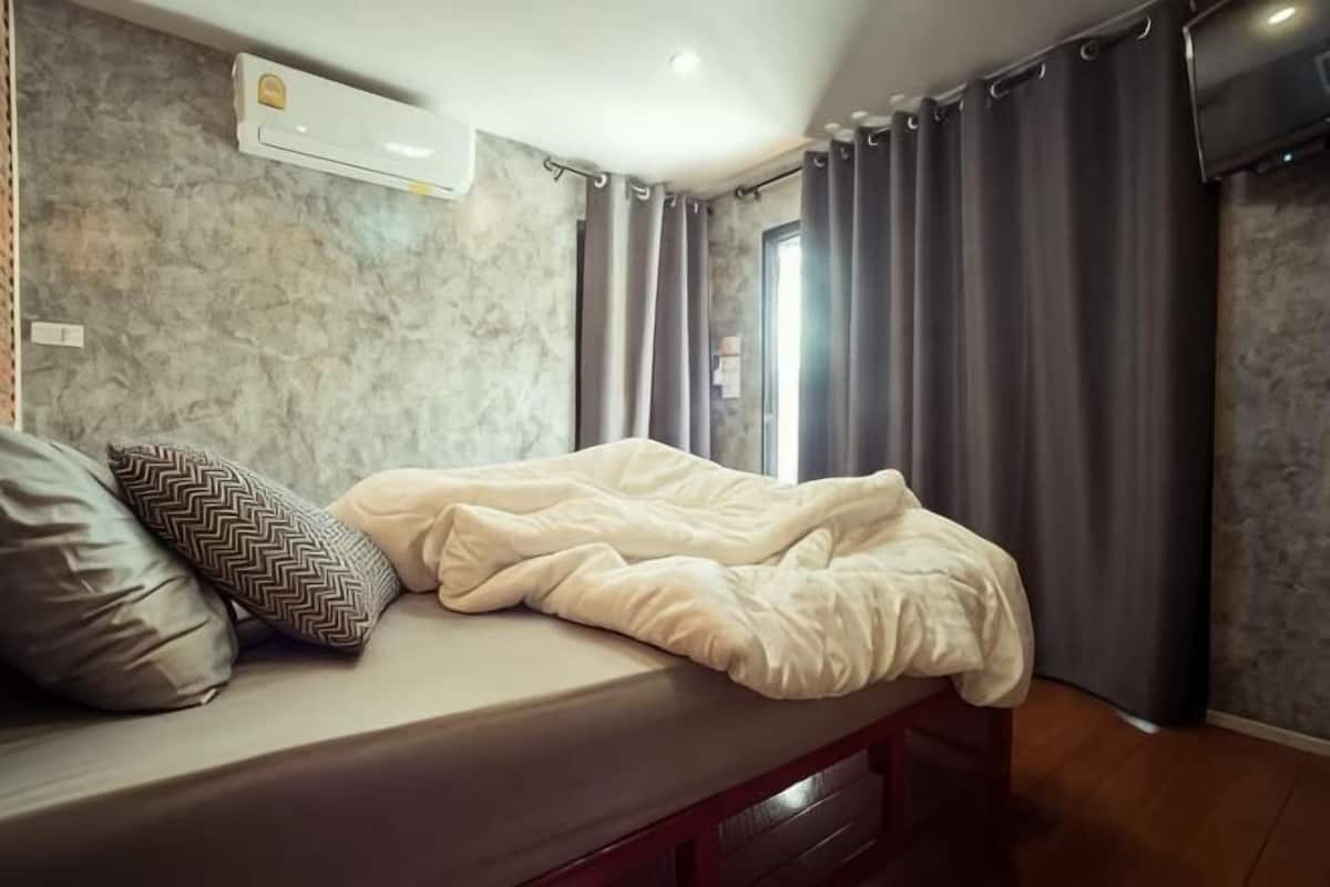 Uma cama com edredom branco e almofadas, parede de mármore, ar condicionado no teto e cortinas cinzas.
