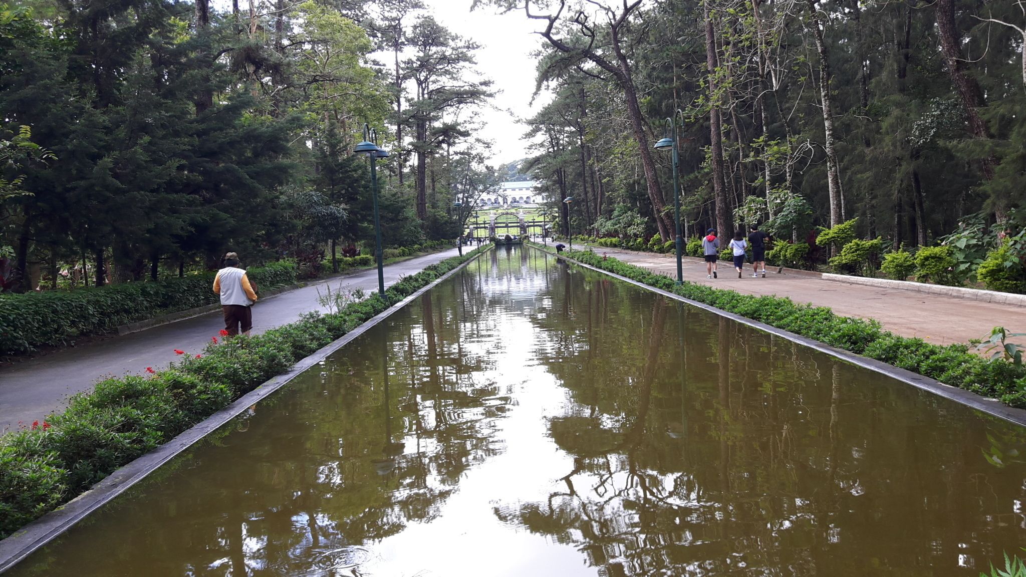 Um canal comprido e estreito ladeado por árvores e uma passarela com pessoas caminhando.