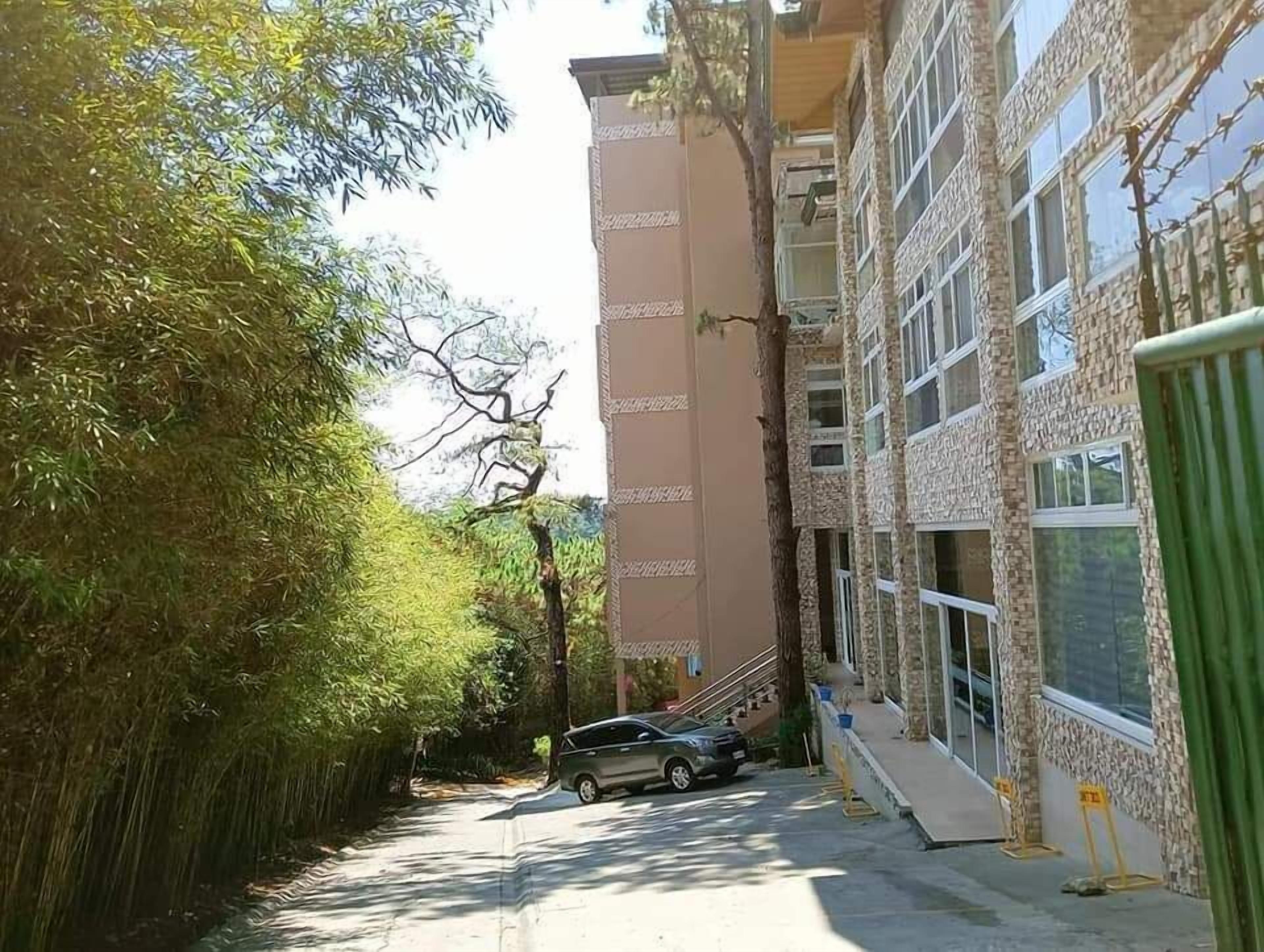 A narrow street with a parked car, a building with a stone facade, and a tree-lined path.