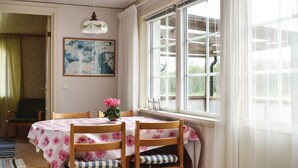 Comedor con una mesa cubierta con mantel floral, dos sillas de madera con cojines a rayas, una lámpara colgante y un ventanal con vistas a la vegetación.