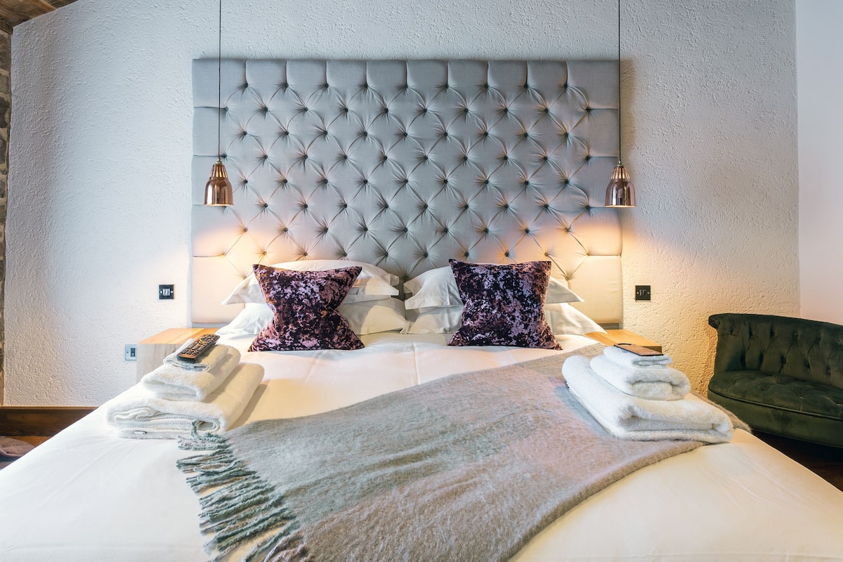 A neatly made bed with a tufted headboard, two bedside lamps, and a green armchair.
