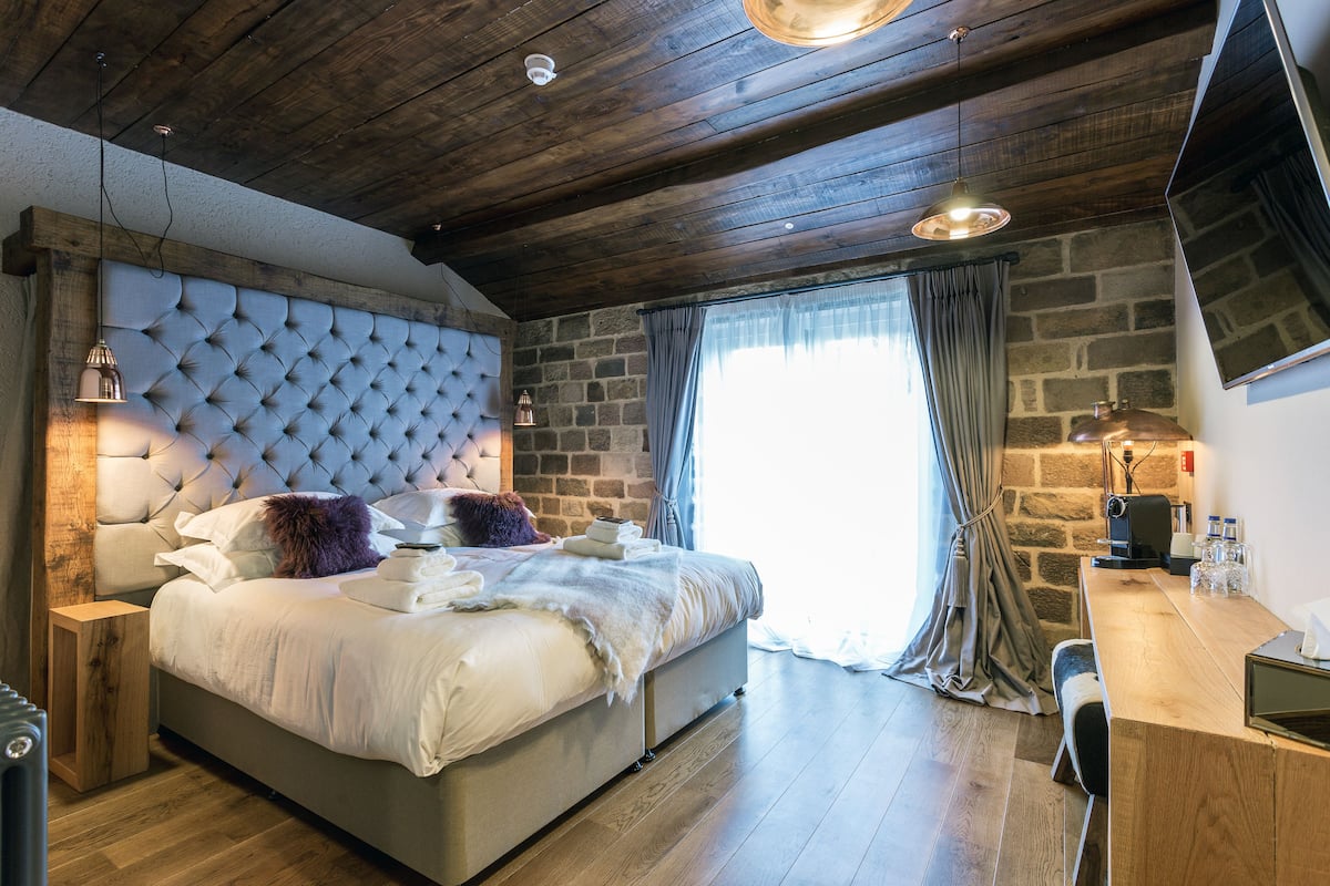 A bedroom with a large bed, a wooden headboard, a stone wall, a wooden desk, and a flat-screen TV.