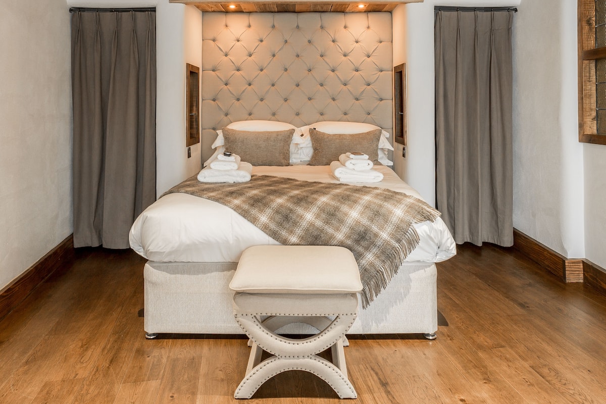 A large bed with a tufted headboard, a bench at the foot of the bed, and grey curtains on either side.