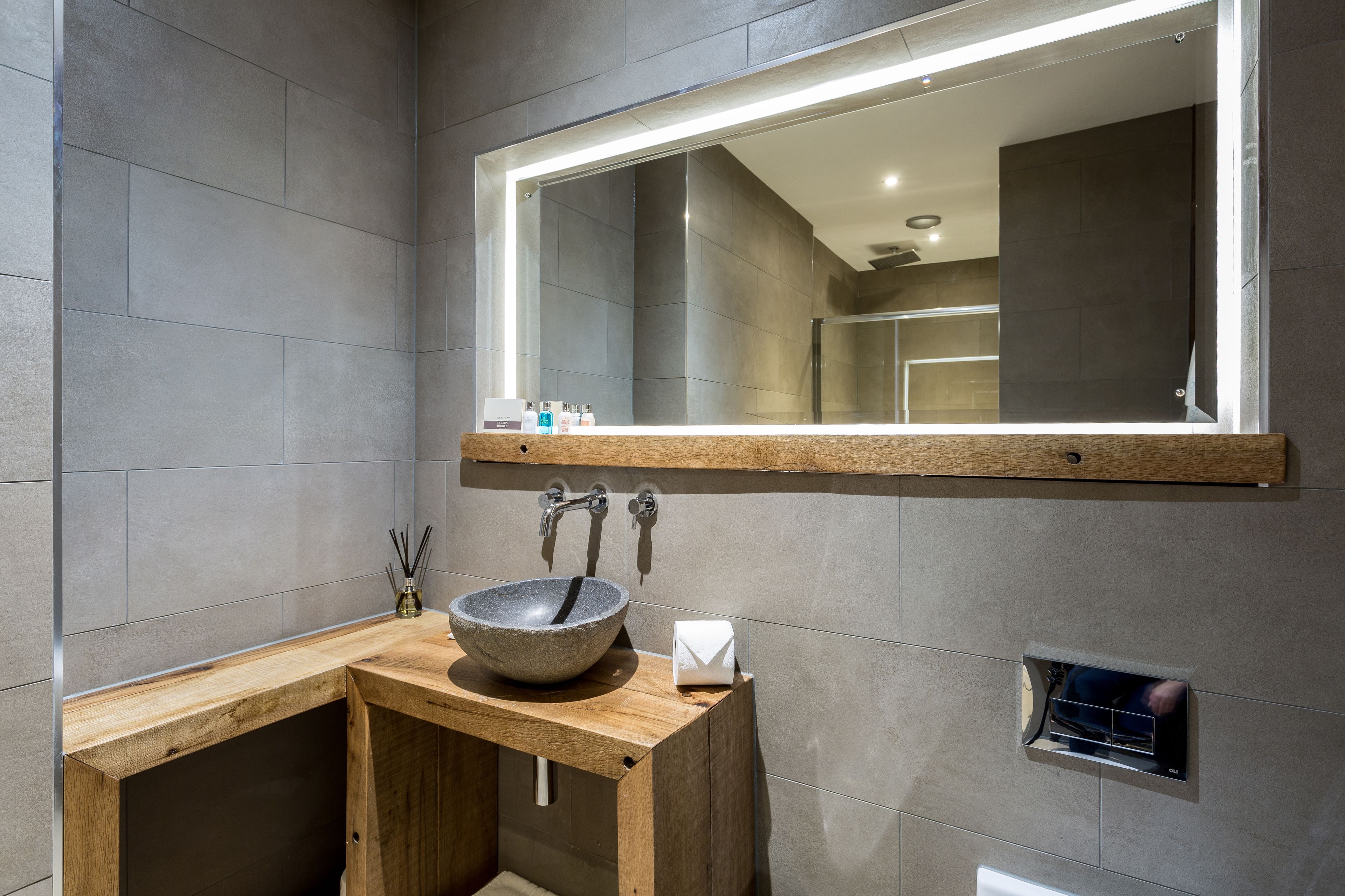 A modern bathroom with a stone sink, wooden vanity, and a large mirror with integrated lighting.