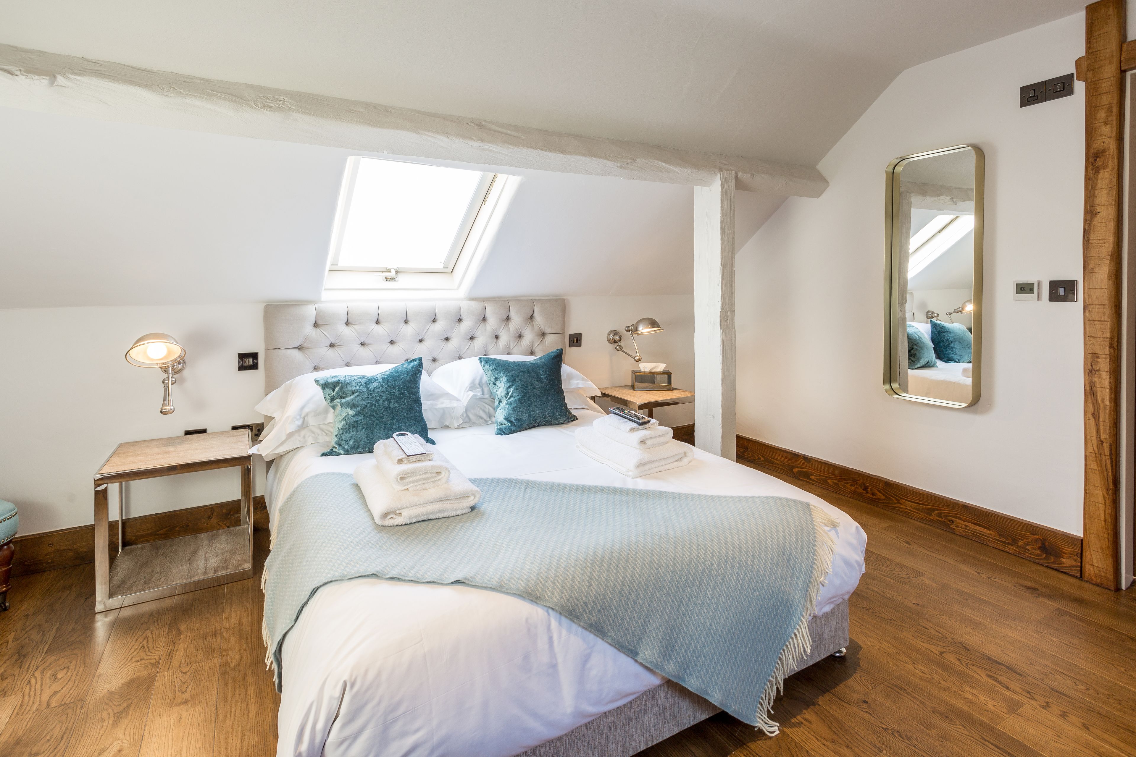 A bedroom with a large bed, a skylight, a mirror, and wooden flooring.