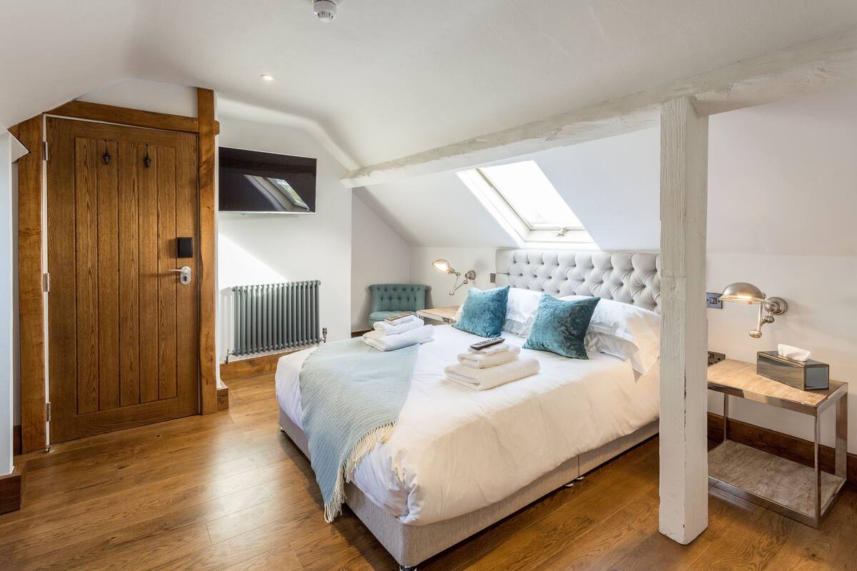 A bedroom with a large bed, wooden flooring, a skylight, a radiator, and a bedside table with a lamp.