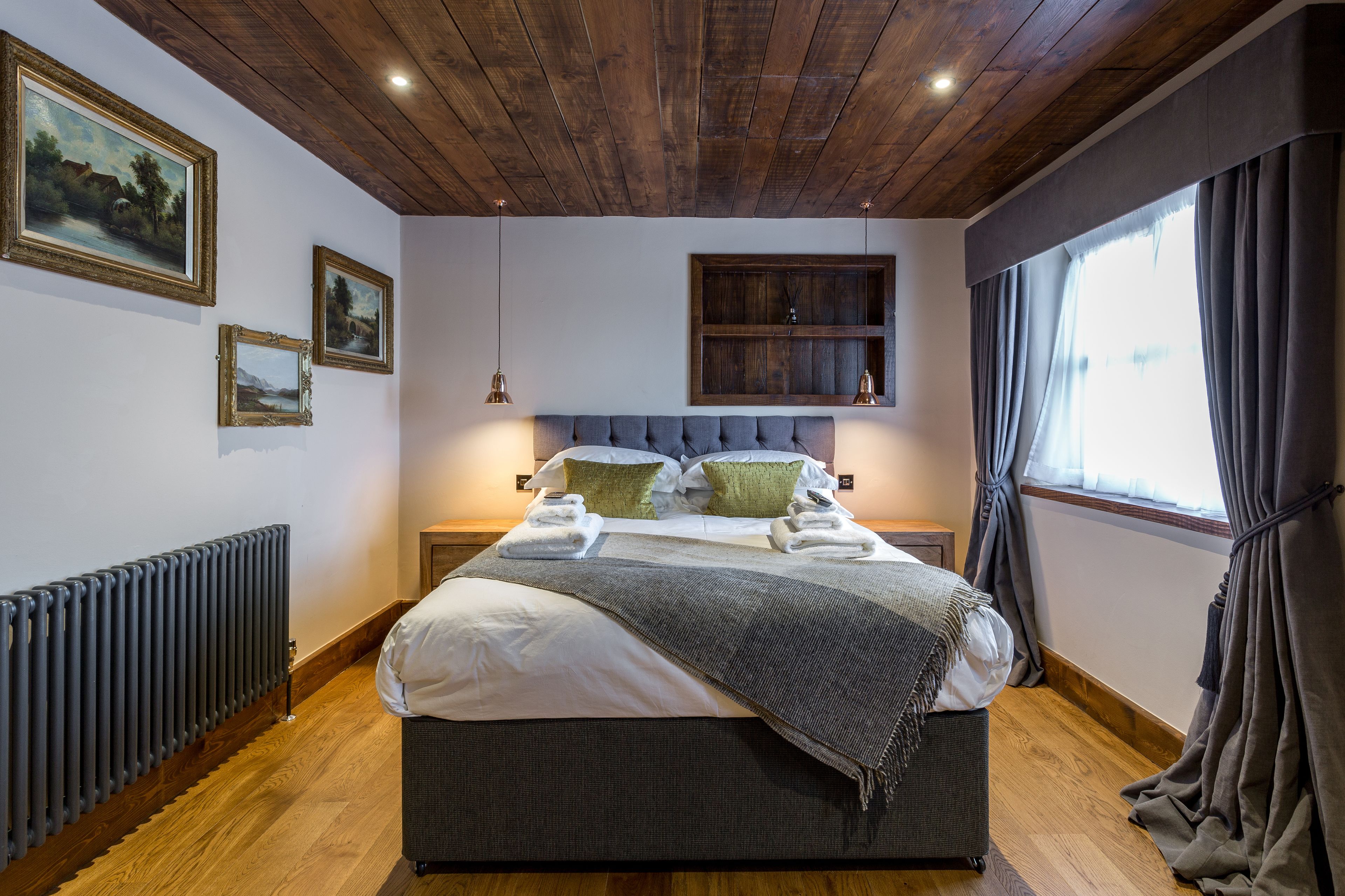 Un dormitorio con techo de madera, una cama grande con almohadas y una colcha, un radiador y cuadros enmarcados en la pared.