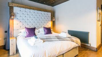 A neatly made bed with a tufted headboard, two purple pillows, and a blanket. There is a wooden nightstand with a lamp and a remote control on it. A radiator is visible on the wall, and the room has wooden flooring.