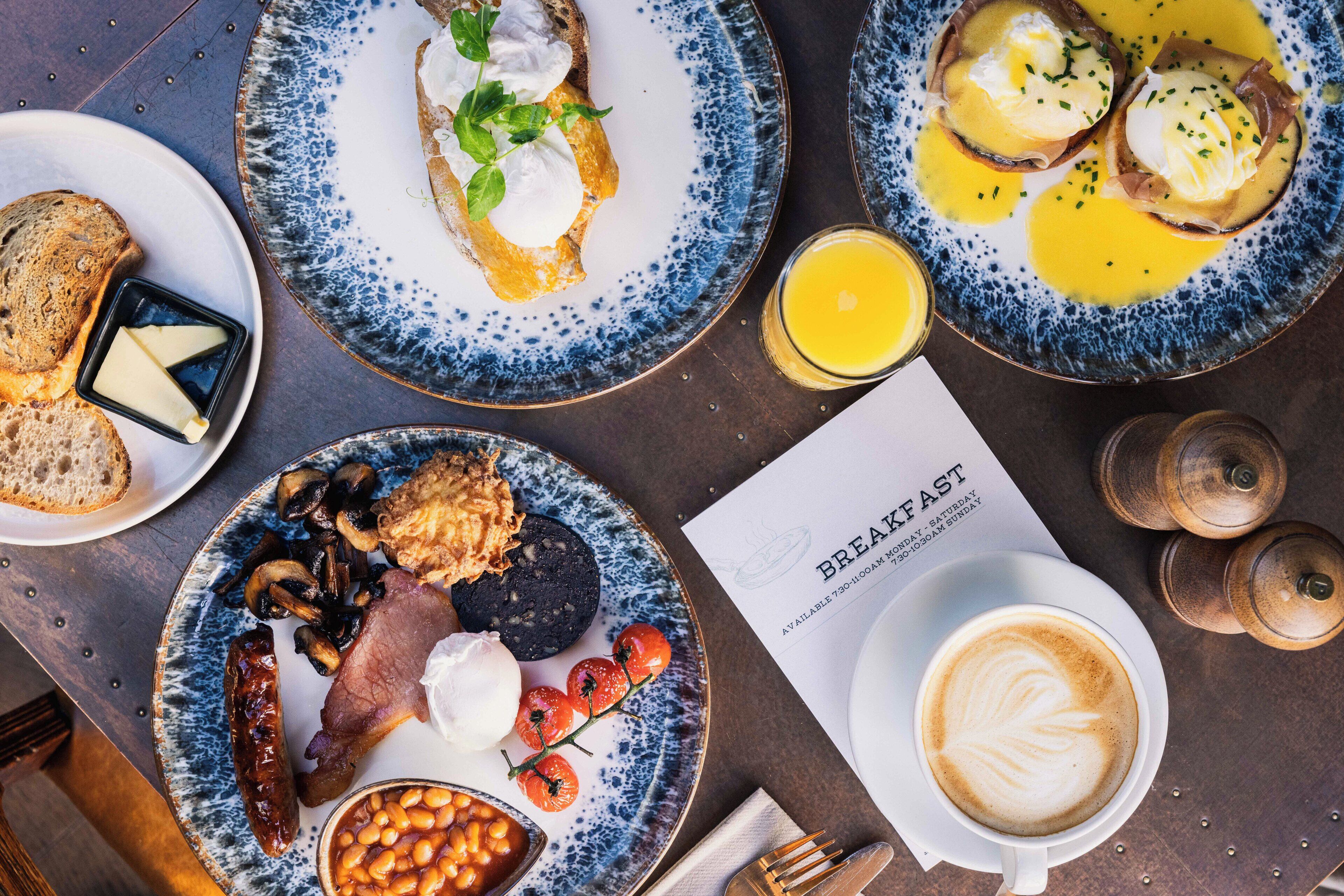 A breakfast setting with plates of eggs, toast, beans, sausages, and a coffee cup.