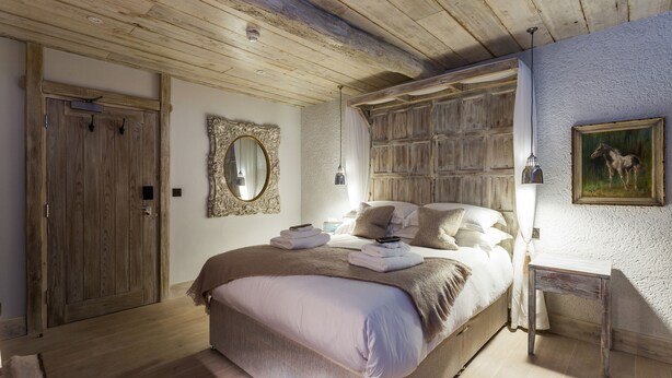 A bedroom with a wooden ceiling, a large bed with a beige blanket, a bedside table with a lamp, and a painting on the wall. - The Cow Dalbury (Ashbourne)