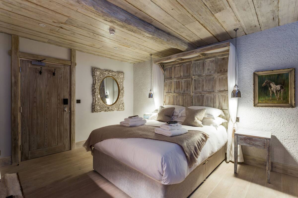 A bedroom with a wooden ceiling, a large bed with a beige blanket, a bedside table with a lamp, and a painting on the wall.