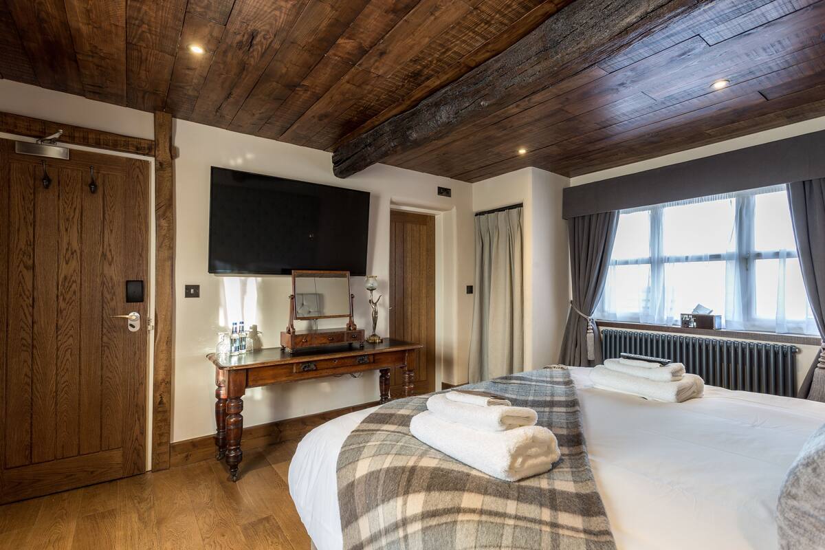 A hotel room with a wooden ceiling, a large bed with a plaid bedspread, a bedside table with a mirror, a dresser, and a window with curtains.