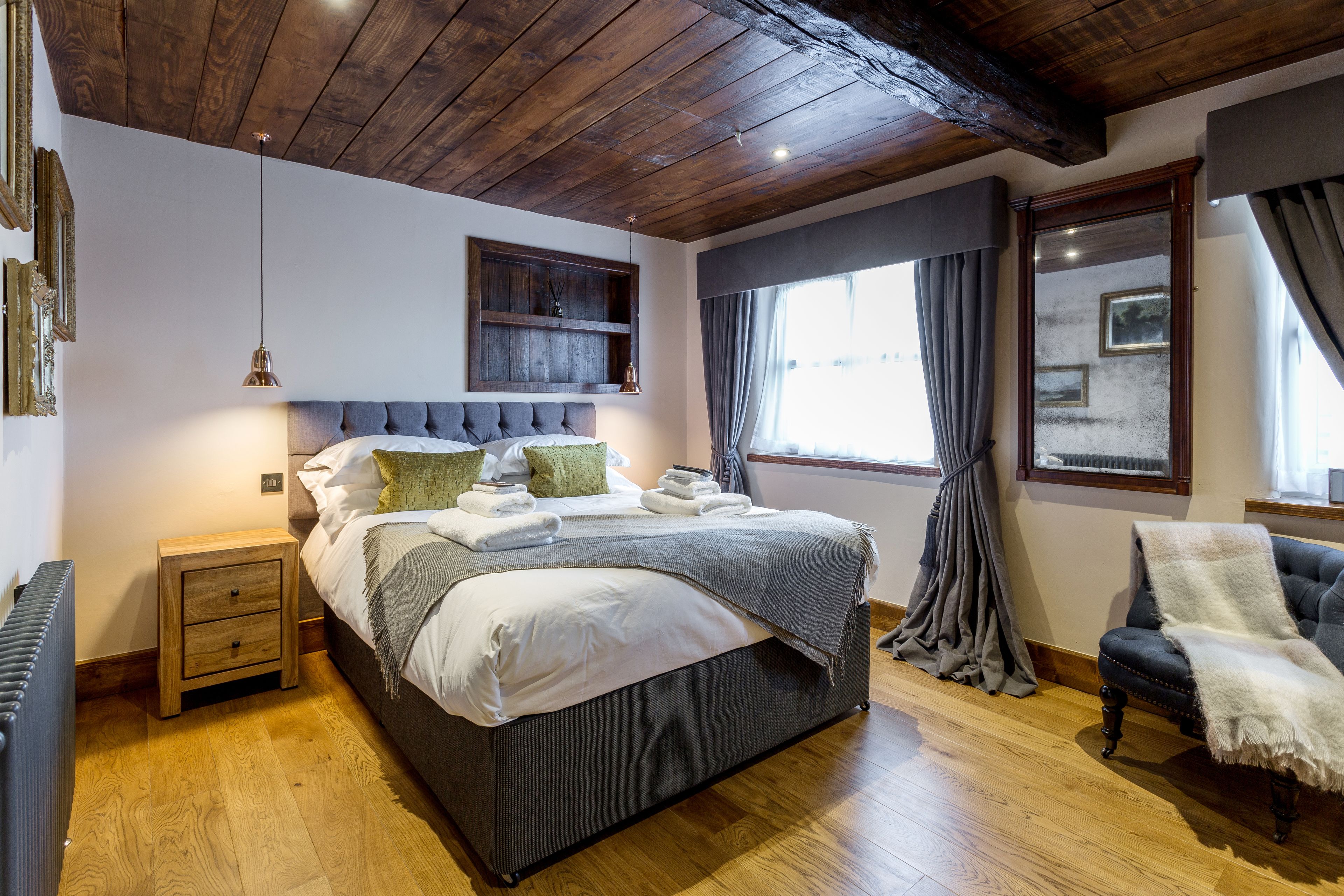 A well-lit bedroom with a large bed, wooden flooring, a nightstand, a chair, and a window with curtains.