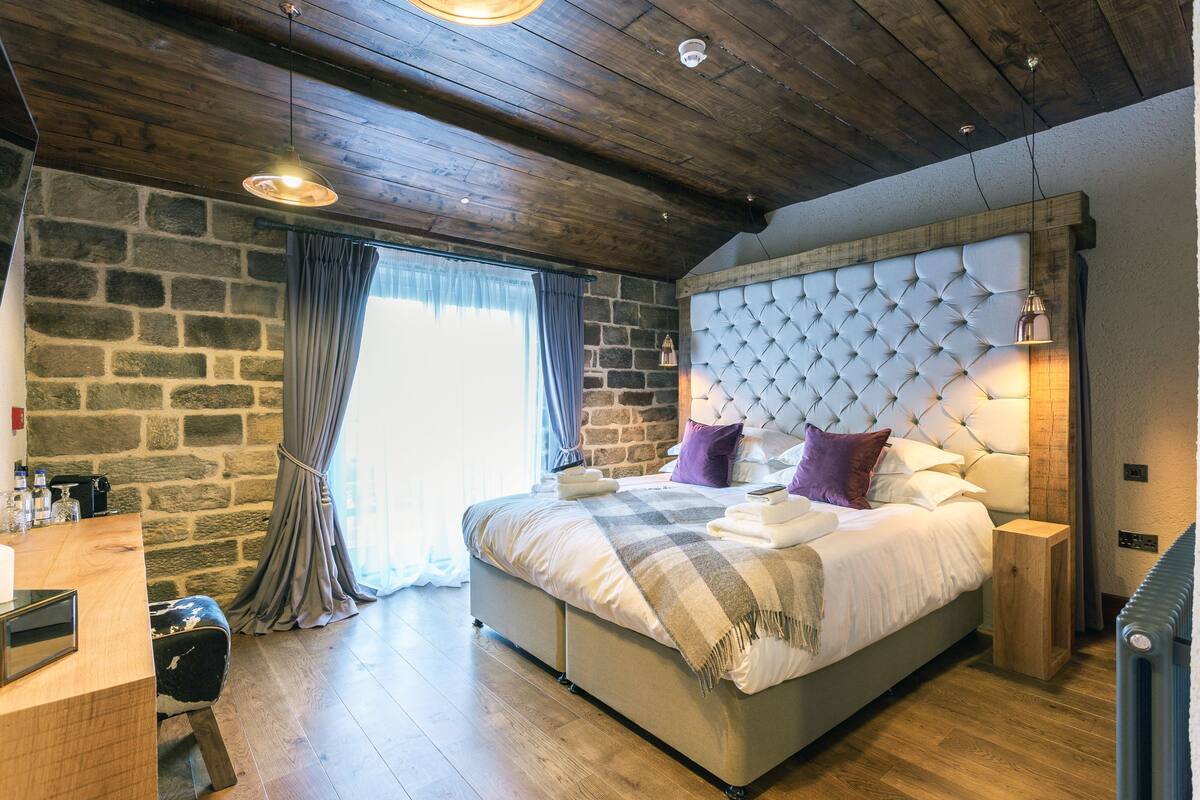 A bedroom with a stone wall, a wooden ceiling, a large bed with a tufted headboard, and a small wooden side table.
