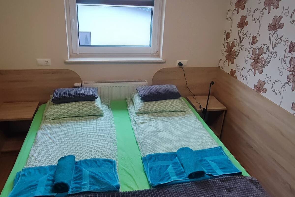 Un lit simple avec une couvre-lit à motifs verts et blancs, deux oreillers et une serviette bleue. Il y a une fenêtre avec des stores, un radiateur en dessous de la fenêtre et un papier peint à motifs floraux sur le mur.