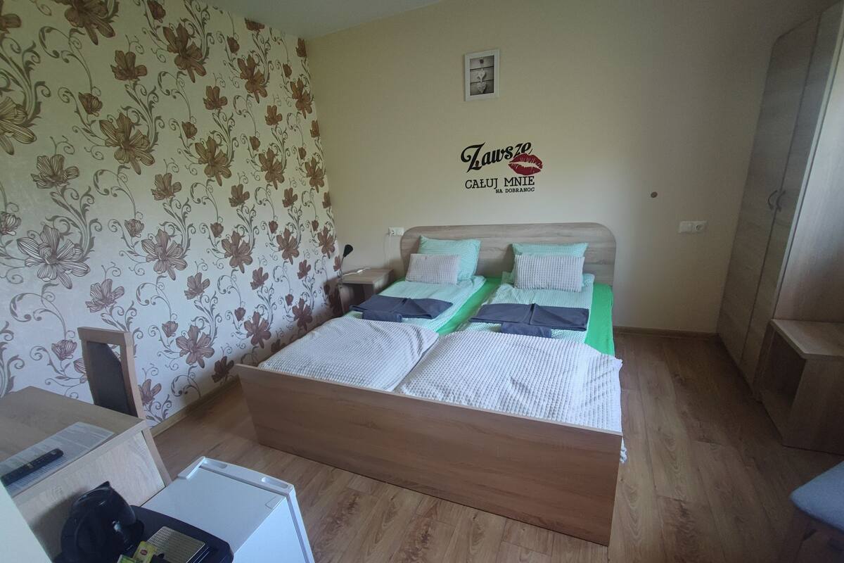 Une chambre à coucher avec un lit en bois, une literie verte et blanche, une petite table de chevet et un mur orné d’un motif floral.