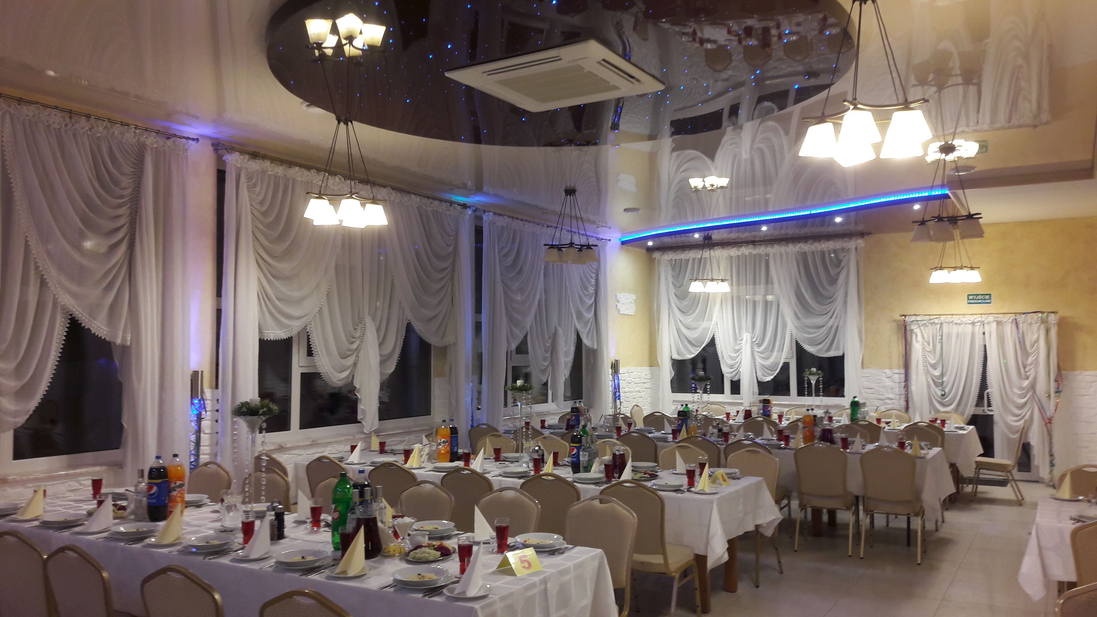 A banquet hall with multiple tables set for dining, large windows with draped curtains, and modern lighting fixtures.