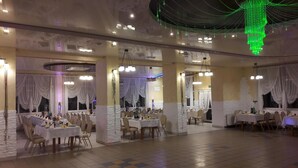 Une salle de banquet spacieuse, dotée de nombreuses tables rondes, de tentures blanches et de luminaires verts suspendus.