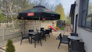 Une terrasse extérieure avec des chaises et des tables en osier noir, un parasol noir et un espace de jeux pour enfants avec un toboggan.