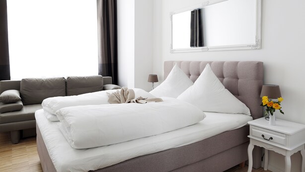 A neatly made bed with white linens, a grey sofa, a bedside table with a lamp, and a mirror on the wall. - SC_3 box spring beds +70 sqm + 3 rooms + balcony + elevator (Berlin)