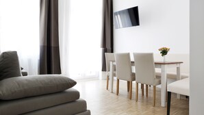 A modern living room with a sofa, a dining table with chairs, a flat-screen TV mounted on the wall, and a window with curtains. - SC_3 box spring beds +70 sqm + 3 rooms + balcony + elevator (Berlin)