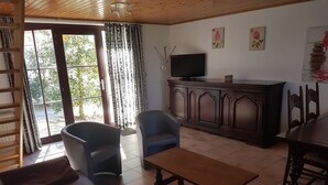 Een woonkamer met een bank, twee stoelen, een houten salontafel, een televisie op een kast en een groot raam met gordijnen.