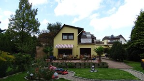 Ein zweistöckiges Gebäude mit gelber Fassade, einer Terrasse mit Sitzgelegenheiten und einem gepflegten Garten.
