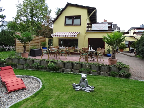 Un edificio de dos pisos con fachada amarilla, un patio cubierto con muebles de mimbre y un pequeño jardín con una estatua de un sapo.