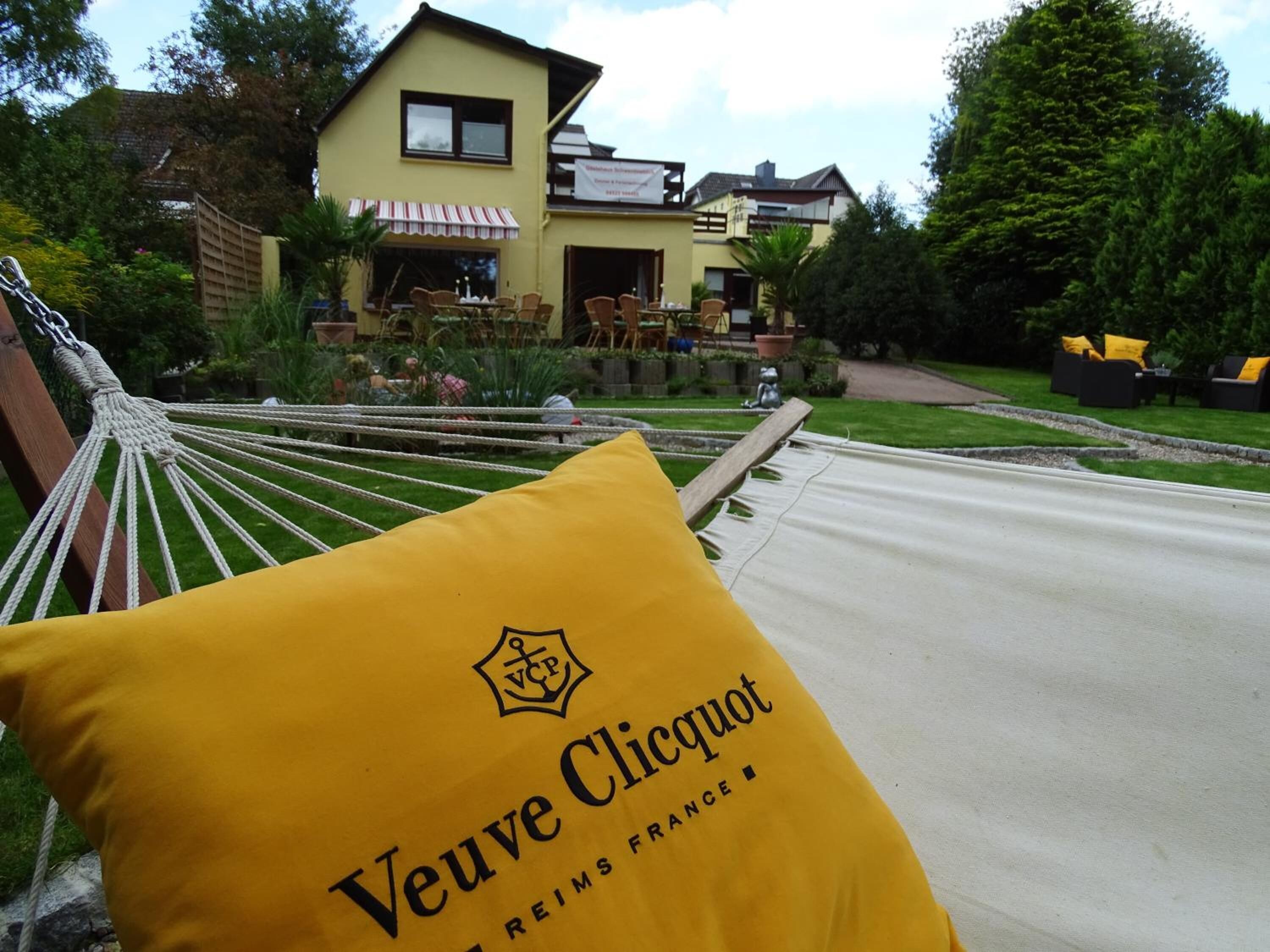 A hammock with a yellow pillow labeled "Veuve Clicquot" in front of a house with a garden.