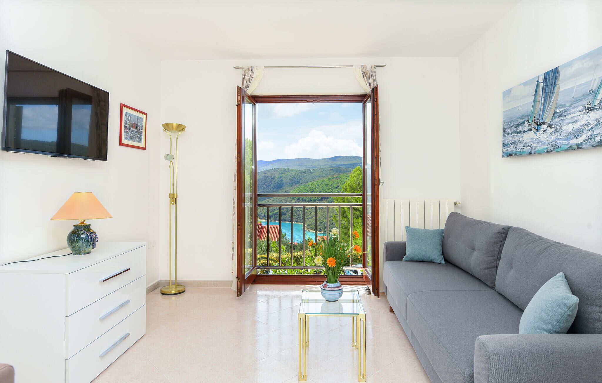 A living room with a sofa, a TV, a lamp, a glass coffee table with flowers, and a balcony with a view of the sea and hills.
