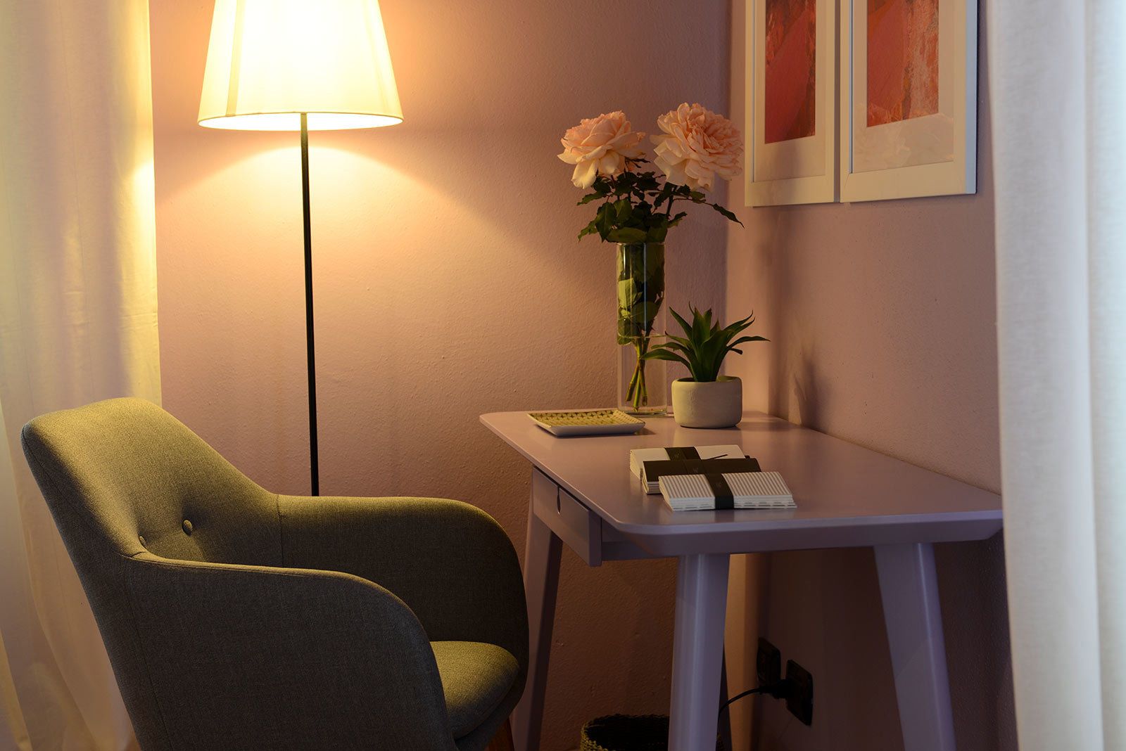Una habitación de esquina con un sillón verde, una mesita con un florero, una lámpara y cuadros en la pared.