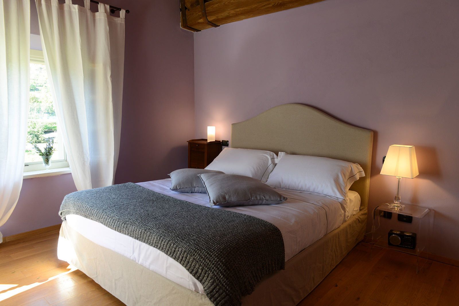 Ein ordentlich bezogenes Bett mit Kissen und Decke, ein Nachttisch mit Lampe und ein Fenster mit Vorhängen.