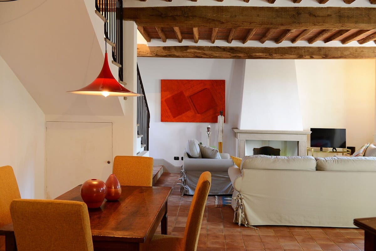 Una sala de estar acogedora con chimenea, un comedor con mesa y sillas de madera, y una escalera con una lámpara colgante roja.